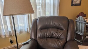 Een bruine leren fauteuil in een kamer met een lamp, een klein tafeltje en een lijst met een afbeelding aan de muur.
