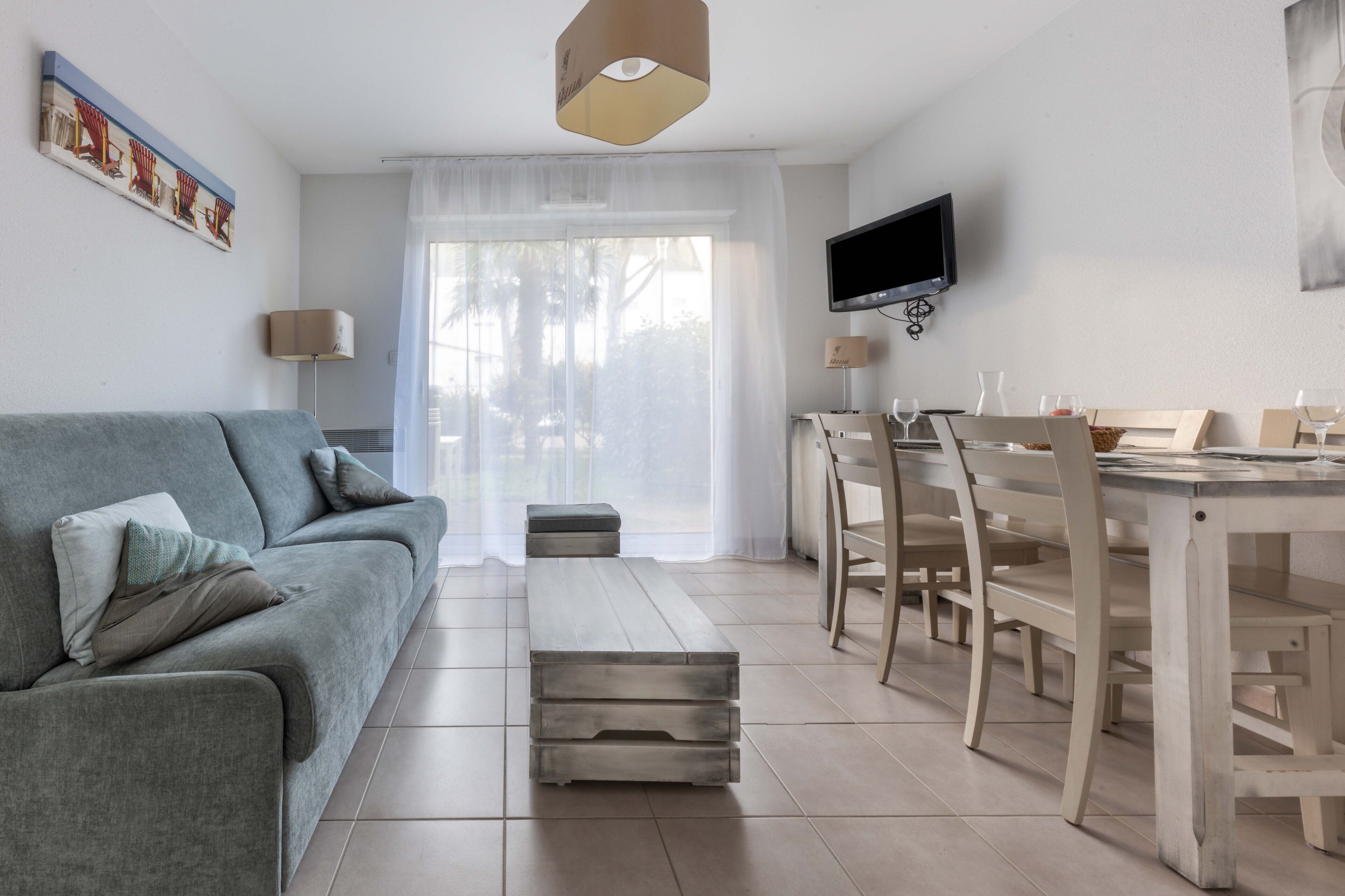 A modern living room with a grey sofa, a wooden dining table with chairs, a flat-screen TV mounted on the wall, and a large window with sheer curtains.