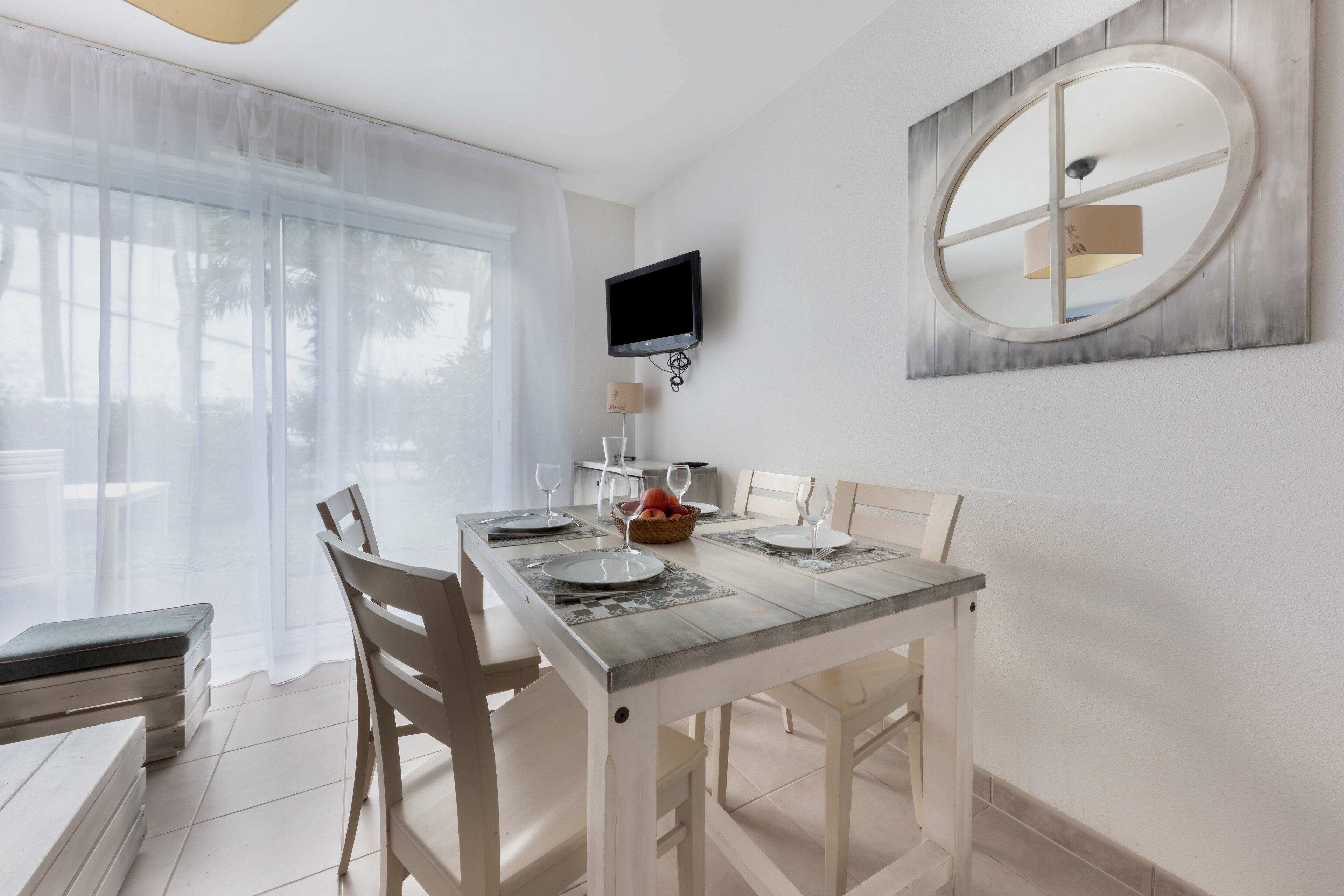 Un coin repas avec une table dressée pour deux personnes, un téléviseur fixé au mur et une fenêtre ornée de rideaux légers.