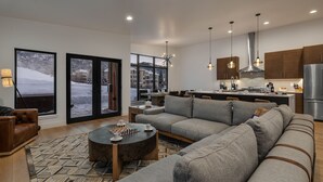 A modern living room with a large sectional sofa, a coffee table, and a chess set. There are large windows with a view of a snowy landscape. The kitchen has wooden cabinets and stainless steel appliances.