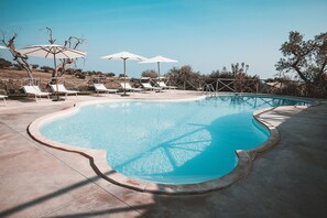A swimming pool with lounge chairs and umbrellas, surrounded by trees and a clear sky.