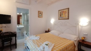 Ein Hotelzimmer mit einem Bett, Nachttisch und Schreibtisch mit Stuhl. An der Wand ist ein Fernseher angebracht, das Badezimmer verfügt über ein Waschbecken und einen Spiegel, und eine Tür führt in einen weiteren Raum.