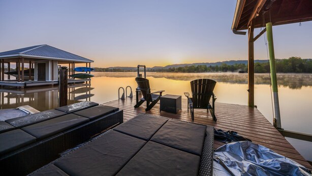 Une terrasse en bois avec des chaises longues et un espace abrité donnant sur un lac paisible au coucher du soleil.