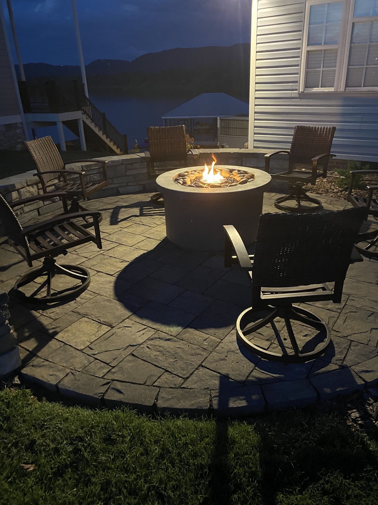 An outdoor patio with a stone floor, a circular fire pit with flames, and wicker chairs.