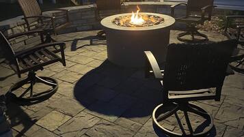 An outdoor patio with a stone floor, a circular fire pit with flames, and wicker chairs.