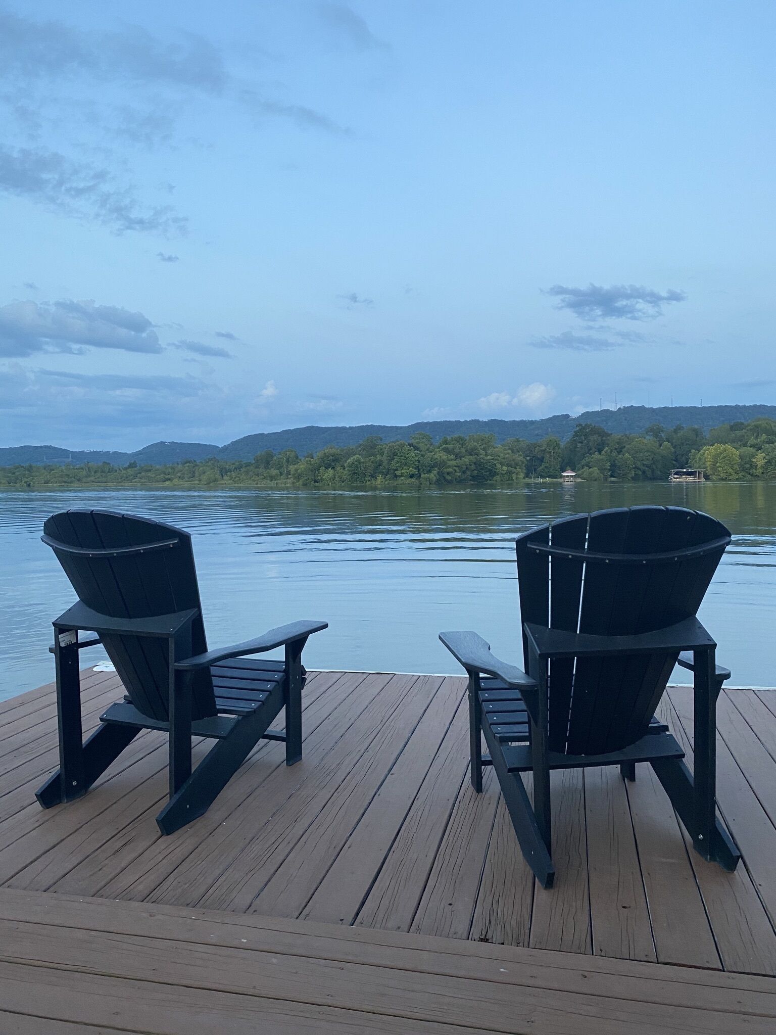 Dos sillas Adirondack en una terraza de madera con vista a un lago, colinas y una construcción pequeña en la distancia.