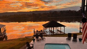 Un espace aménagé au bord de la piscine, avec des chaises longues, un belvédère et une vue sur un lac au coucher du soleil.