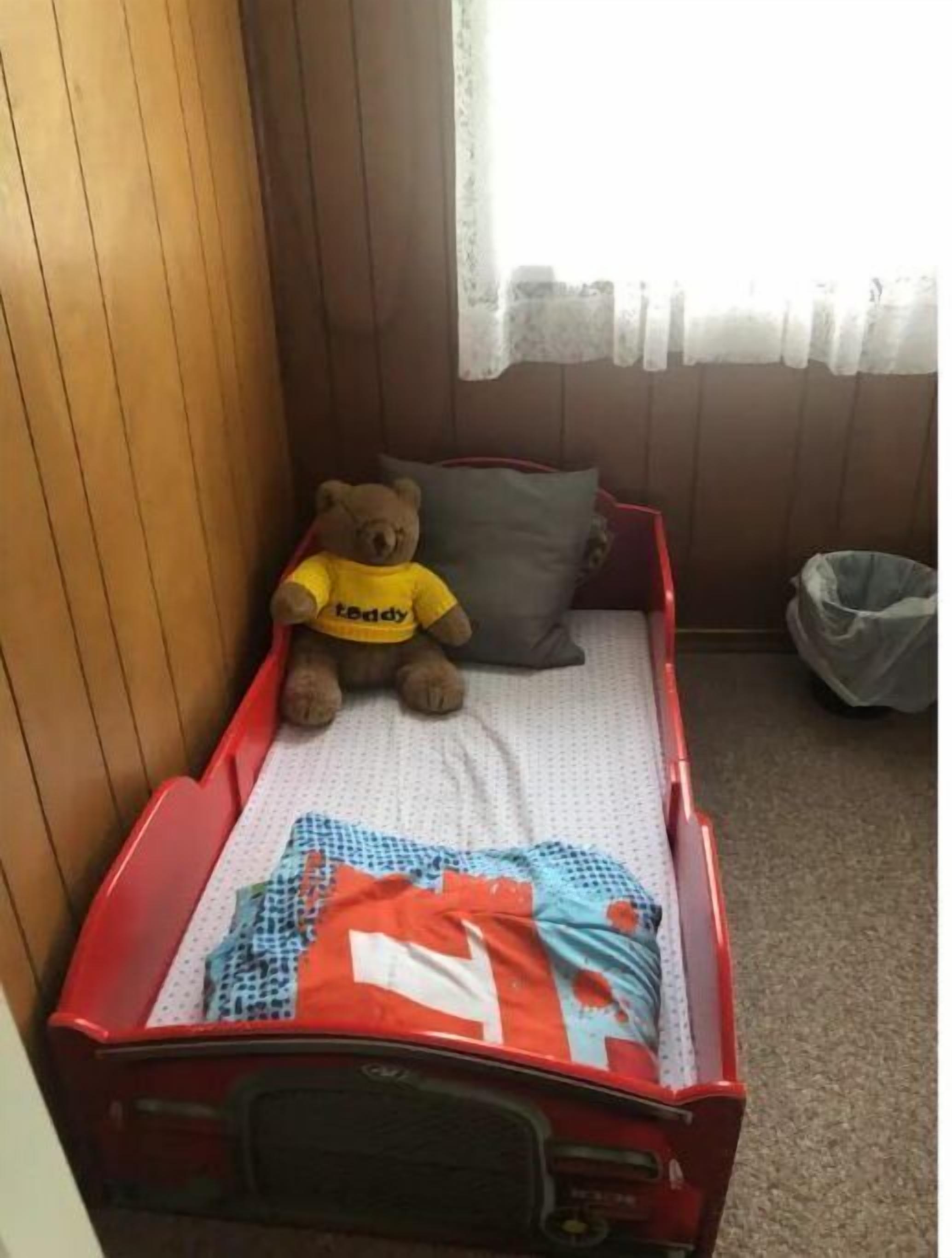Quarto pequeno com uma cama de solteiro, uma cama vermelha, um urso de pelúcia vestindo uma camisa amarela, um travesseiro cinzento e uma cortina branca.