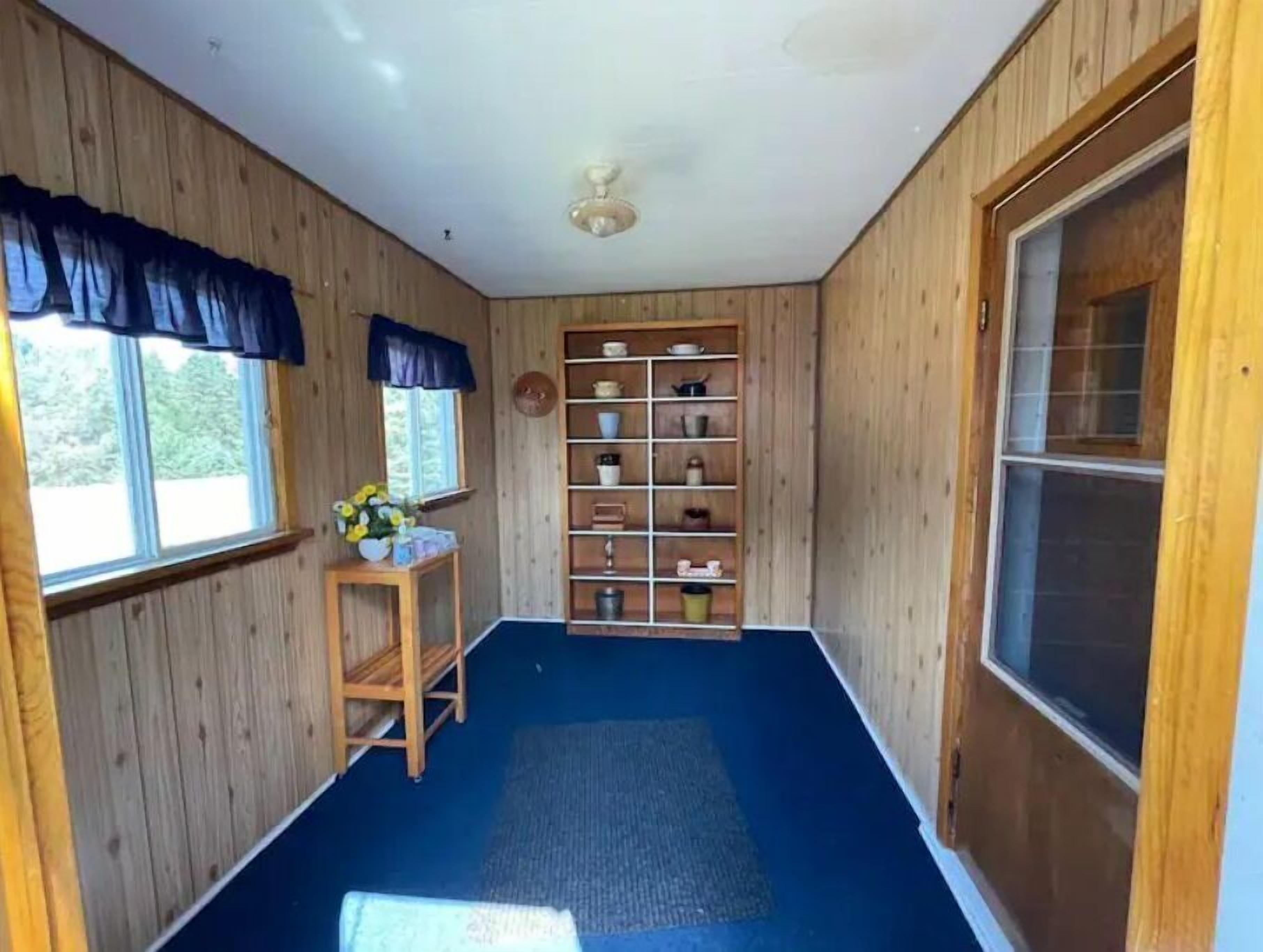 A room with wooden walls, a blue carpet, a ceiling fan, a window with curtains, a shelf with kitchenware, and a door.