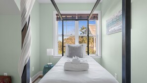 Un lit bien fait, avec du linge de lit blanc et une serviette pliée, une fenêtre donnant sur des palmiers et un tableau accroché au mur.