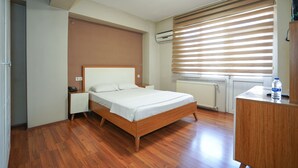 A bedroom with a wooden bed, a bedside table, a nightstand, a radiator, and a window with blinds. - İpek Otel (Balikesir)