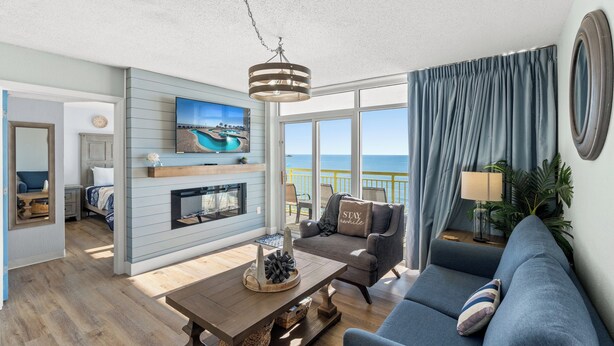 A living room with a sofa, a coffee table, and a fireplace. There is a television mounted on the wall and a window with curtains.