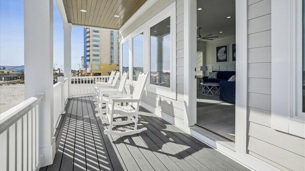 A wooden deck with white rocking chairs, a white railing, and a view of the beach and a tall building in the background. - The Beach Club 8BR OF House in Cherry Grove (North Myrtle Beach)