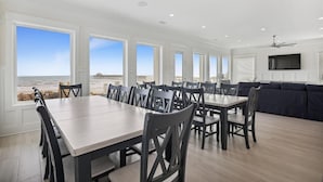A spacious dining area with a long table and chairs, large windows with a view of the beach, a ceiling fan, and a TV mounted on the wall. - The Beach Club 8BR OF House in Cherry Grove (North Myrtle Beach)