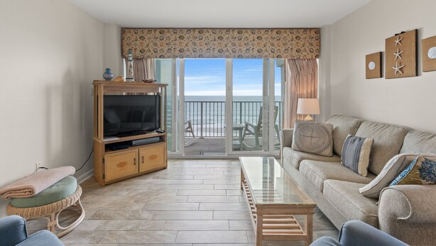A living room with a television, sofa, and a view of the sea through large glass doors. - Carolina Dunes 502 2BR OF Condo in Cherry Grove (North Myrtle Beach)