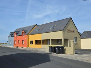 Una hilera de casas coloridas con un letrero que dice 'LE RELAIS DES HUES' y contenedores de basura negros en la entrada.