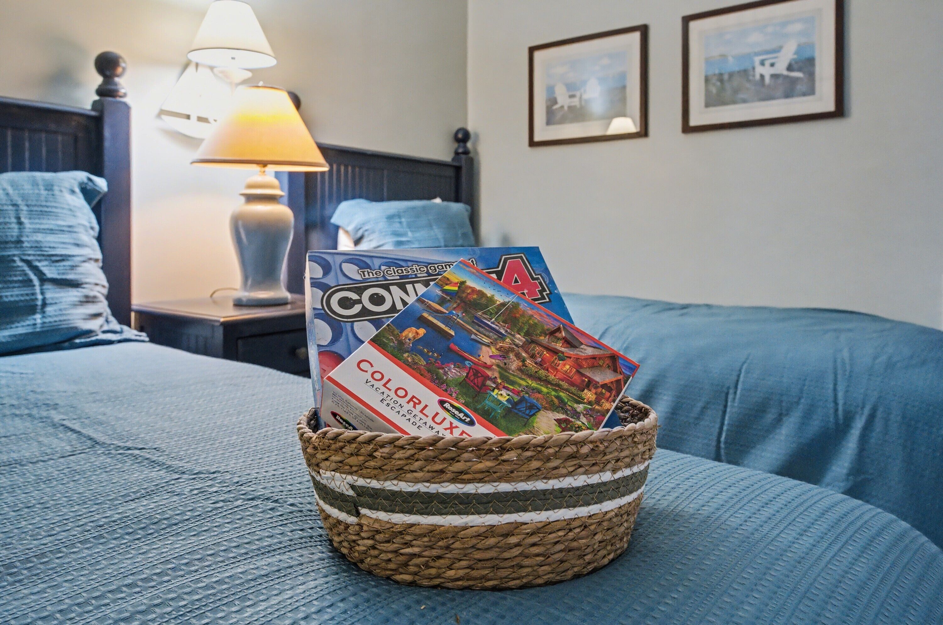 A bed with a woven basket containing board games, a lamp, and framed pictures on the wall.