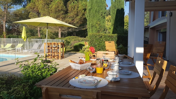 A wooden table set for breakfast with a view of a pool and lounge chairs. - Beautiful architect-designed villa with swimming pool in the middle of a large park 10 minutes from Auch. (Auterive)