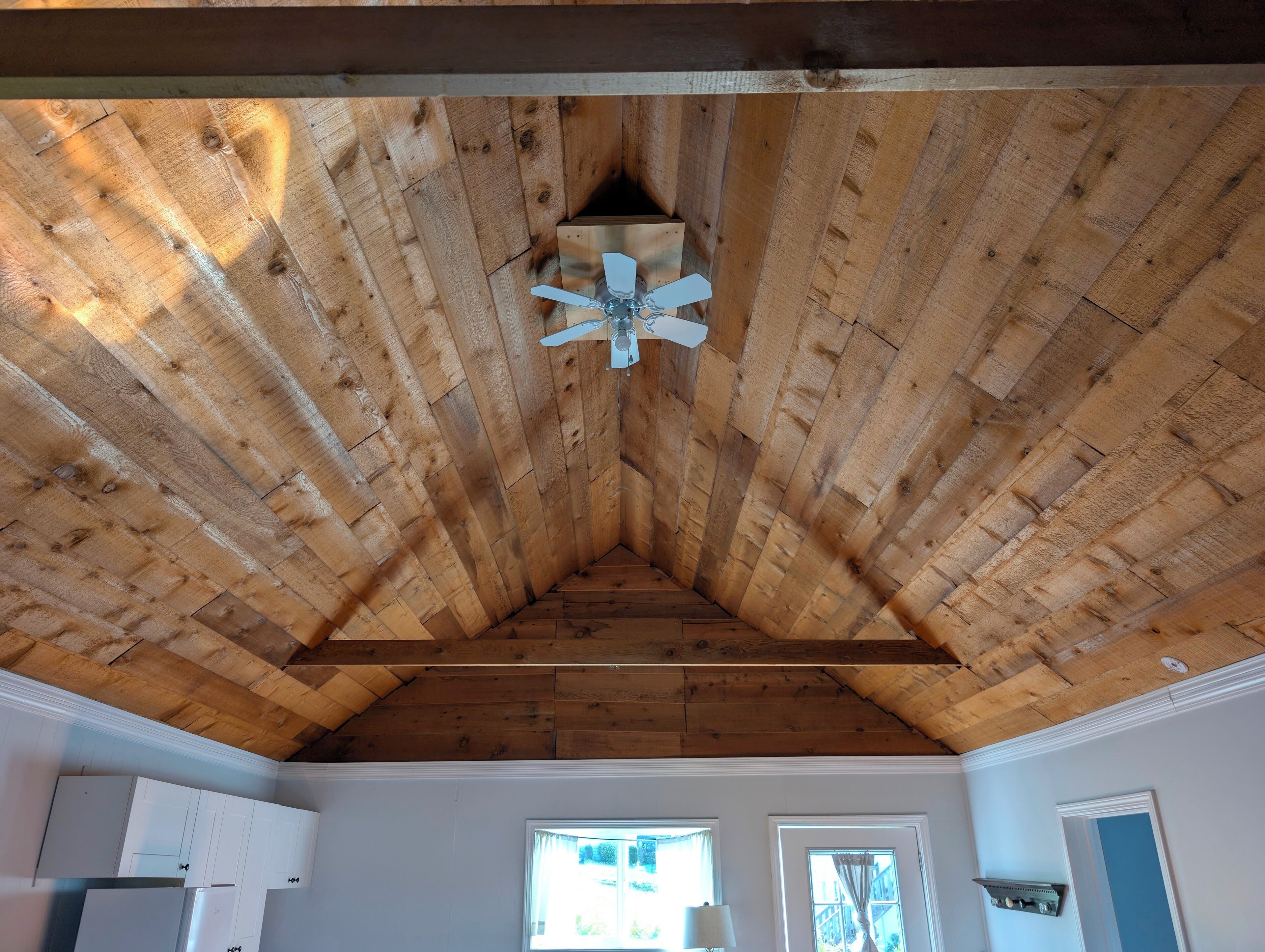 Une pièce avec un plafond en bois incliné, un ventilateur de plafond, des murs blancs et une fenêtre avec des rideaux.