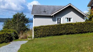A house with a gravel driveway leading to a grassy area, a hedge, and a view of the water. - Cliff Haven on the Bay (Alert Bay)