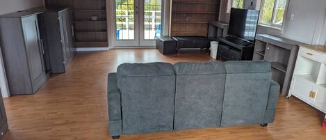 A spacious living area with a grey sofa, wooden flooring, a television, and a sliding glass door leading to a balcony.