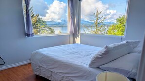 A bedroom with a bed, a window with curtains, and a view of a lake and mountains. - Cliff Haven on the Bay (Alert Bay)