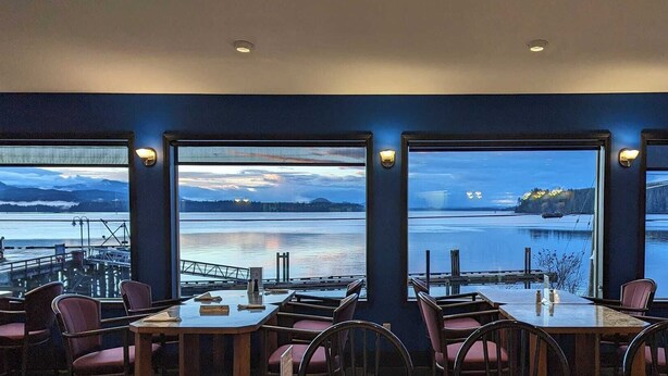 A restaurant with large windows offering views of a lake and mountains. - Cliff Haven on the Bay (Alert Bay)