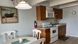 A compact kitchen with white cabinets, a white stove, and a microwave. There is a dining table with white chairs and a teal placemat. A fire extinguisher is mounted on the wall, and a clock is visible on the wall. - Perfect Getaway. Only 1 block to the Beach (Seaside Park)