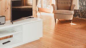 Un salón moderno con paredes de madera, un televisor de pantalla plana, un mueble blanco para equipo audiovisual, un sillón cómodo y un gran ventanal con cortinas.