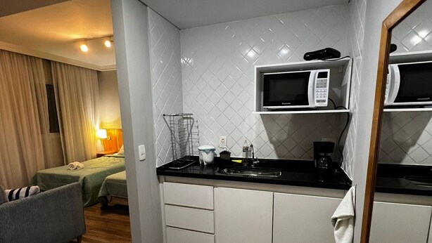 A compact kitchenette with a microwave, stove, and small countertop. There is a bed with a grey headboard and a blue rug in the adjacent room. - Elegance Suites Oscar Freire (São Paulo)