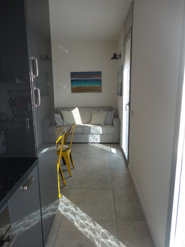 A modern kitchen with a black refrigerator, a dining area with a yellow chair, and a living room with a sofa and a painting.