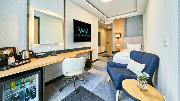 A hotel room with a desk, a chair, a bed, a small table with flowers, and a minibar. - WENS Hotel (Mainz)