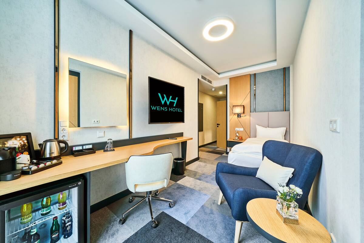 Ein Hotelzimmer mit Schreibtisch, Sessel, Bett, einem kleinen Tisch mit Blumen und einer Minibar.