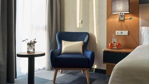 A hotel room with a blue armchair, a small round table with a vase, a bedside table with a lamp, and a bed. - WENS Hotel (Mainz)