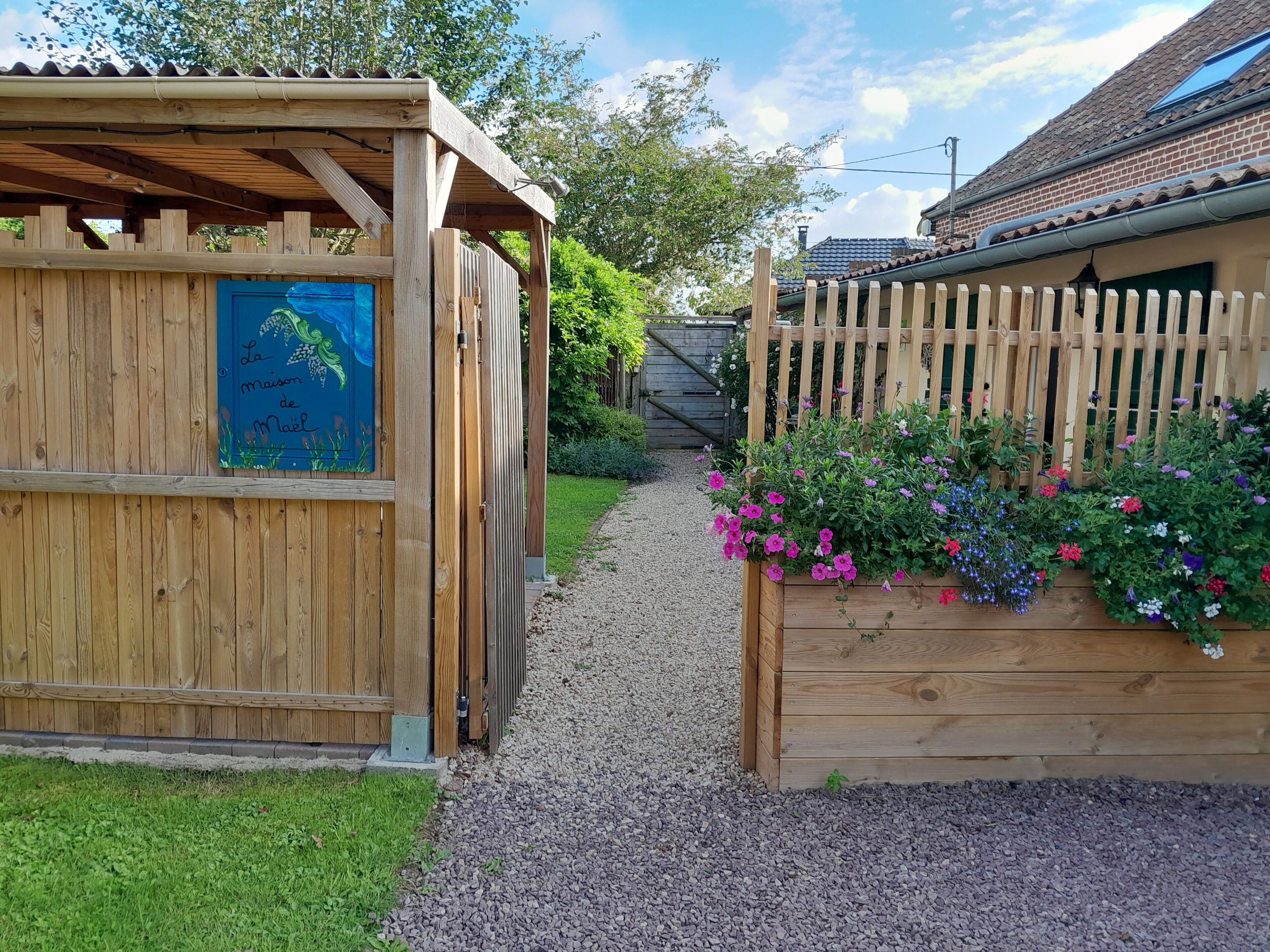 Een houten constructie met een blauw bord, een grindpad en een houten hek met kleurrijke bloemen.