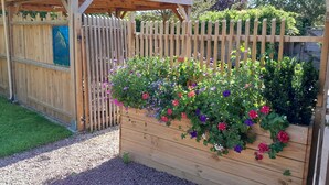 Una estructura de jardín de madera con pérgola, rodeada por una valla de madera y un macizo de flores variado.