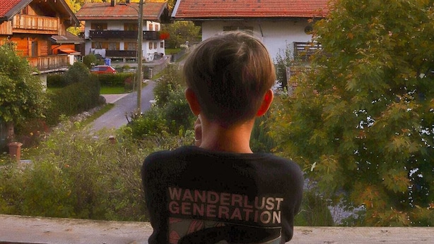 Ein Junge steht auf einem Balkon mit Blick auf ein Dorf, mit Häusern und Bergen im Hintergrund.