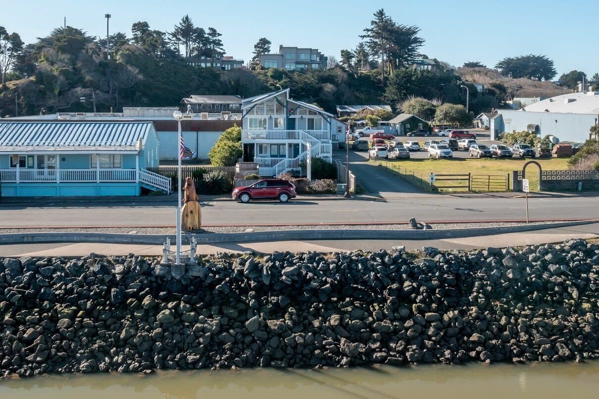 A coastal road with a parking area, a blue building, and a white house with a porch.