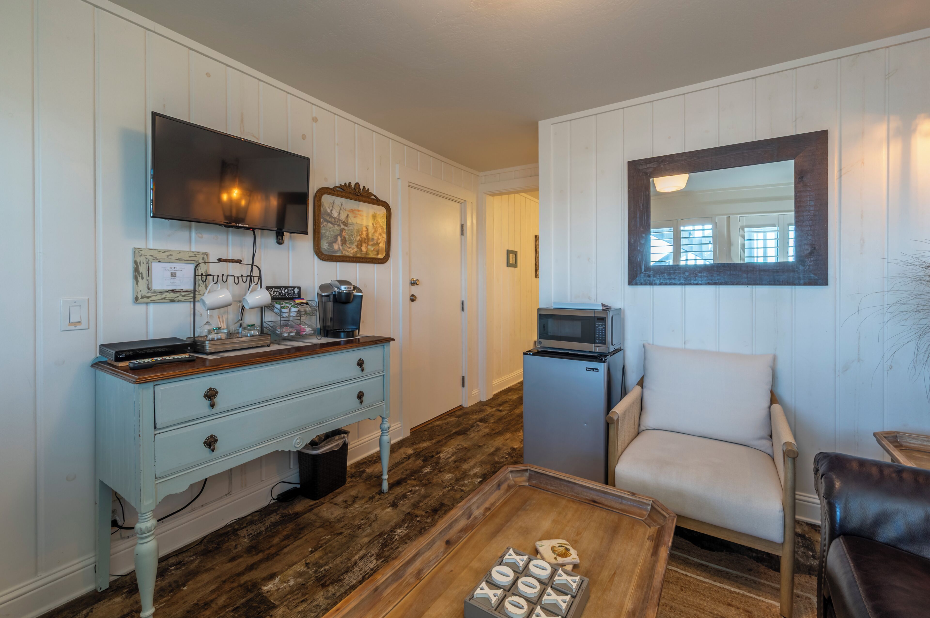 A room with a wooden floor, a light blue dresser, a white armchair, a television, a microwave, and a framed picture on the wall.