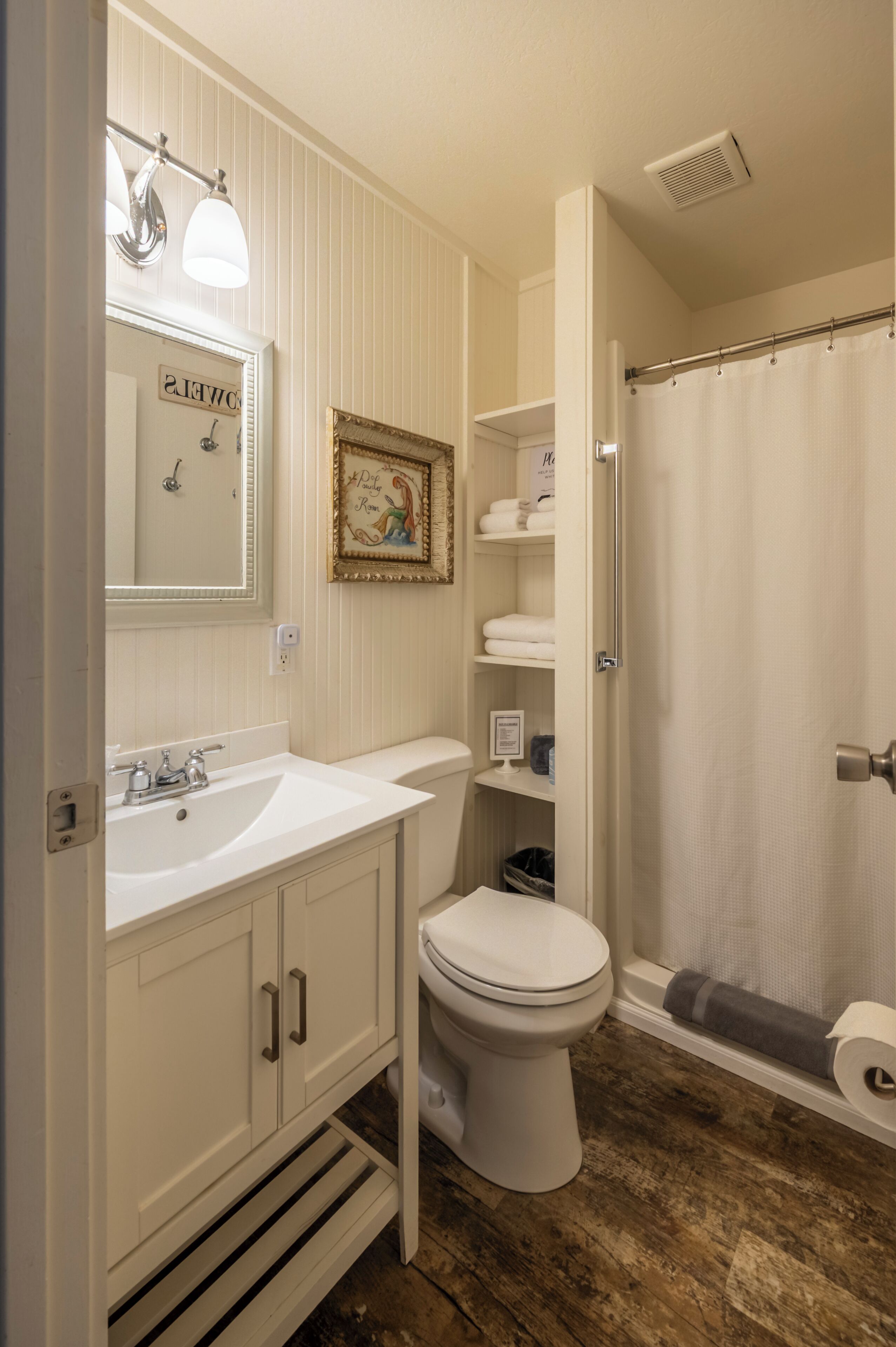 A bathroom with a white sink, toilet, and shower curtain. There is a mirror above the sink and a framed picture on the wall.