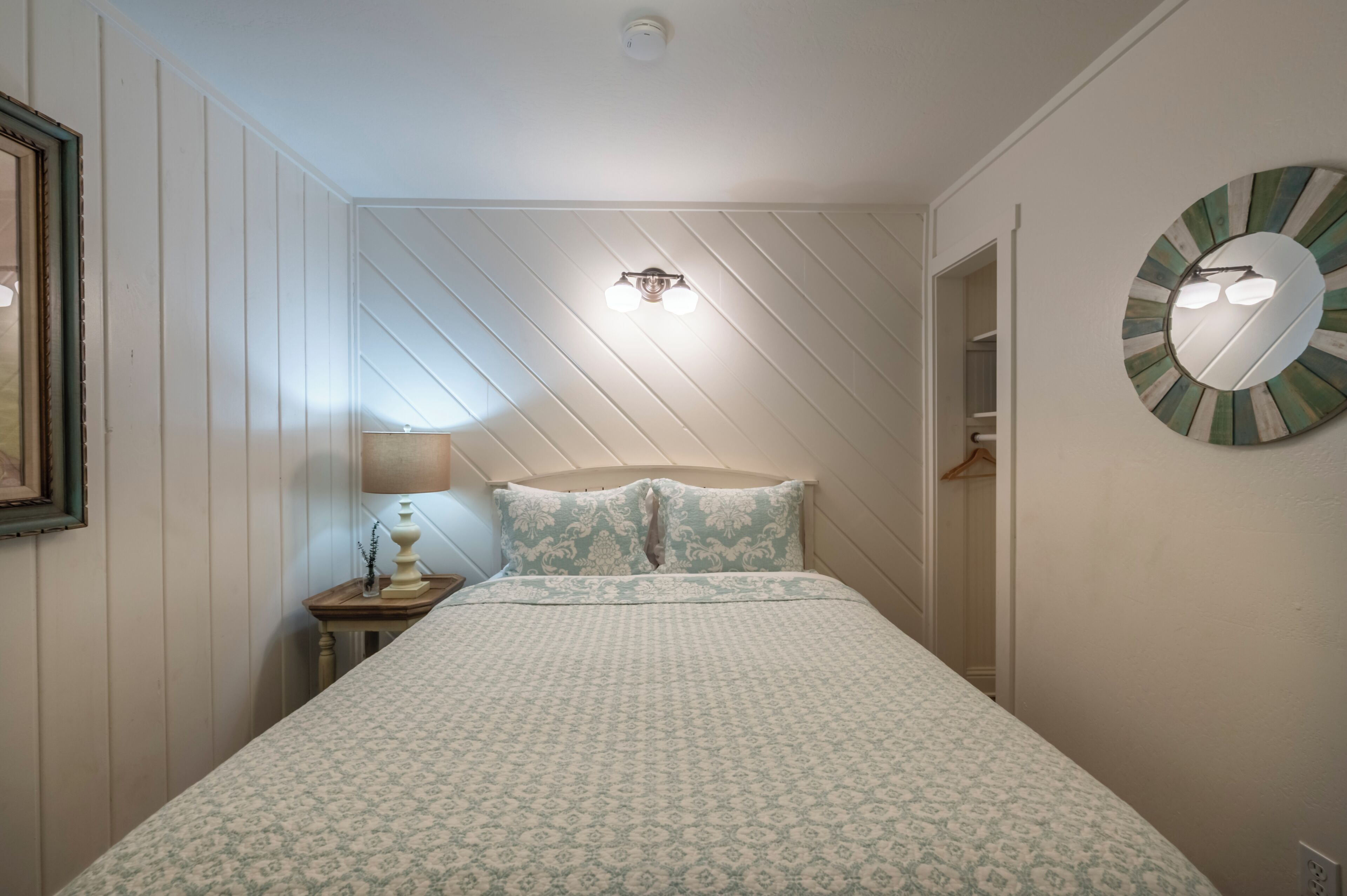 Quarto com cama, mesa de cabeceira, luminária na parede, luminária no teto e espelho redondo.