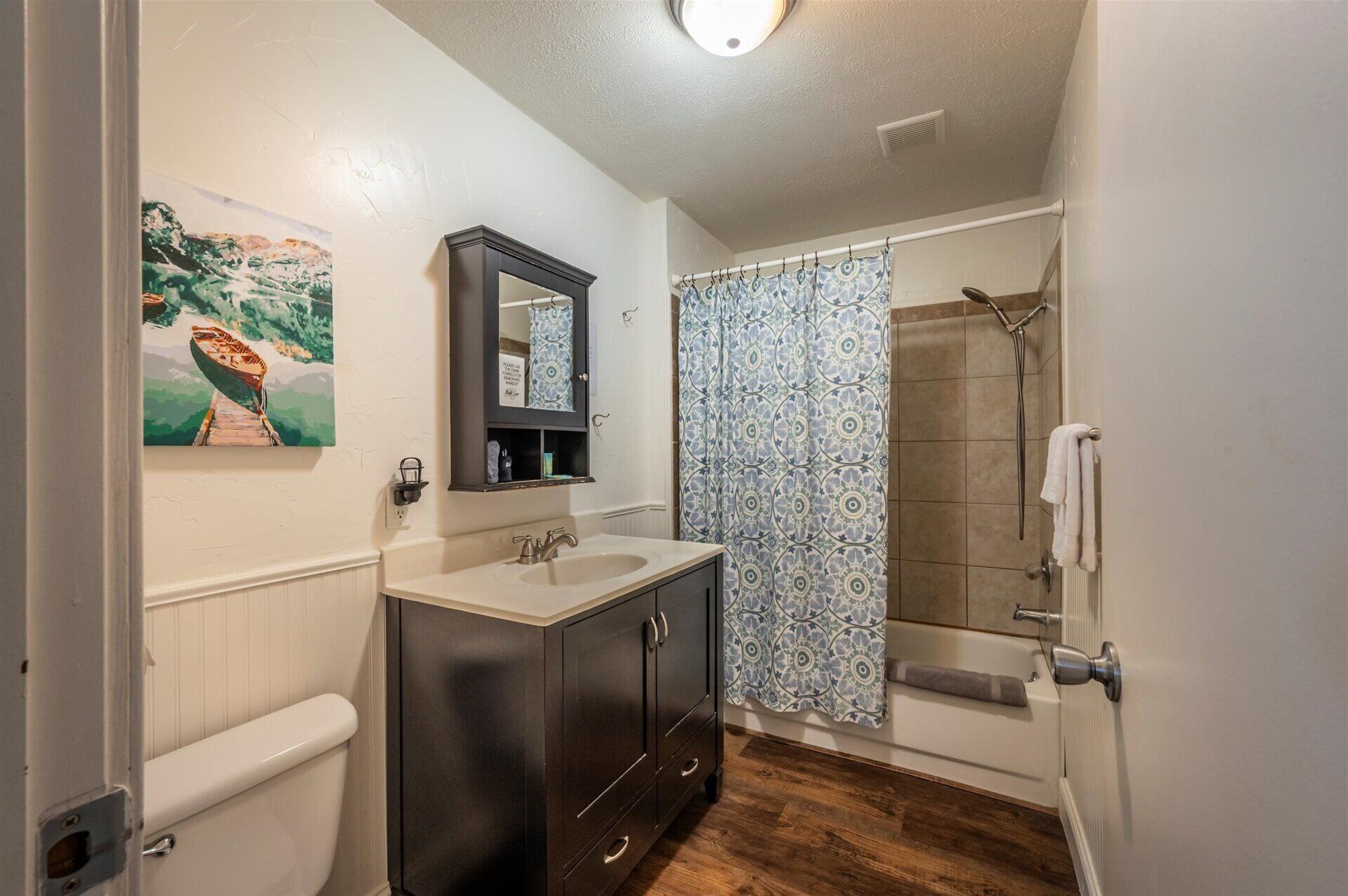 A bathroom with a toilet, dark wooden cabinets, a sink, a mirror, a shower with a patterned curtain, and a painting on the wall.