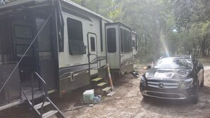 Ein Wohnmobil mit einer Rampe zum Eingang, ein geparktes Auto und ein bewaldeter Hintergrund.