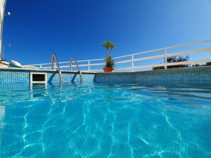 Una piscina azul cristalina con borde de azulejos, una escalera metálica y una planta en maceta al lado.