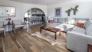 Un salon moderne avec un canapé gris, une table basse en bois et un tapis en peau de vache. On aperçoit à l’arrière-plan un coin repas, avec une cuisine visible à travers une arche.