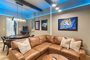 A living room with a brown leather sectional sofa, a coffee table, and a TV mounted on the wall. There are framed posters on the wall and a gaming console in the background.