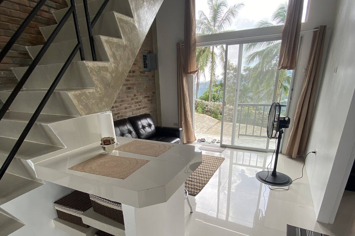 Ruime woonkamer met een moderne trap, een witte bank en een groot raam met uitzicht op palmbomen.