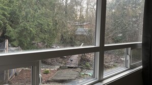 Blick aus einem Fenster auf einen Garten mit einer Holzterrasse, umgeben von Bäumen und einem kleinen Gebäude.