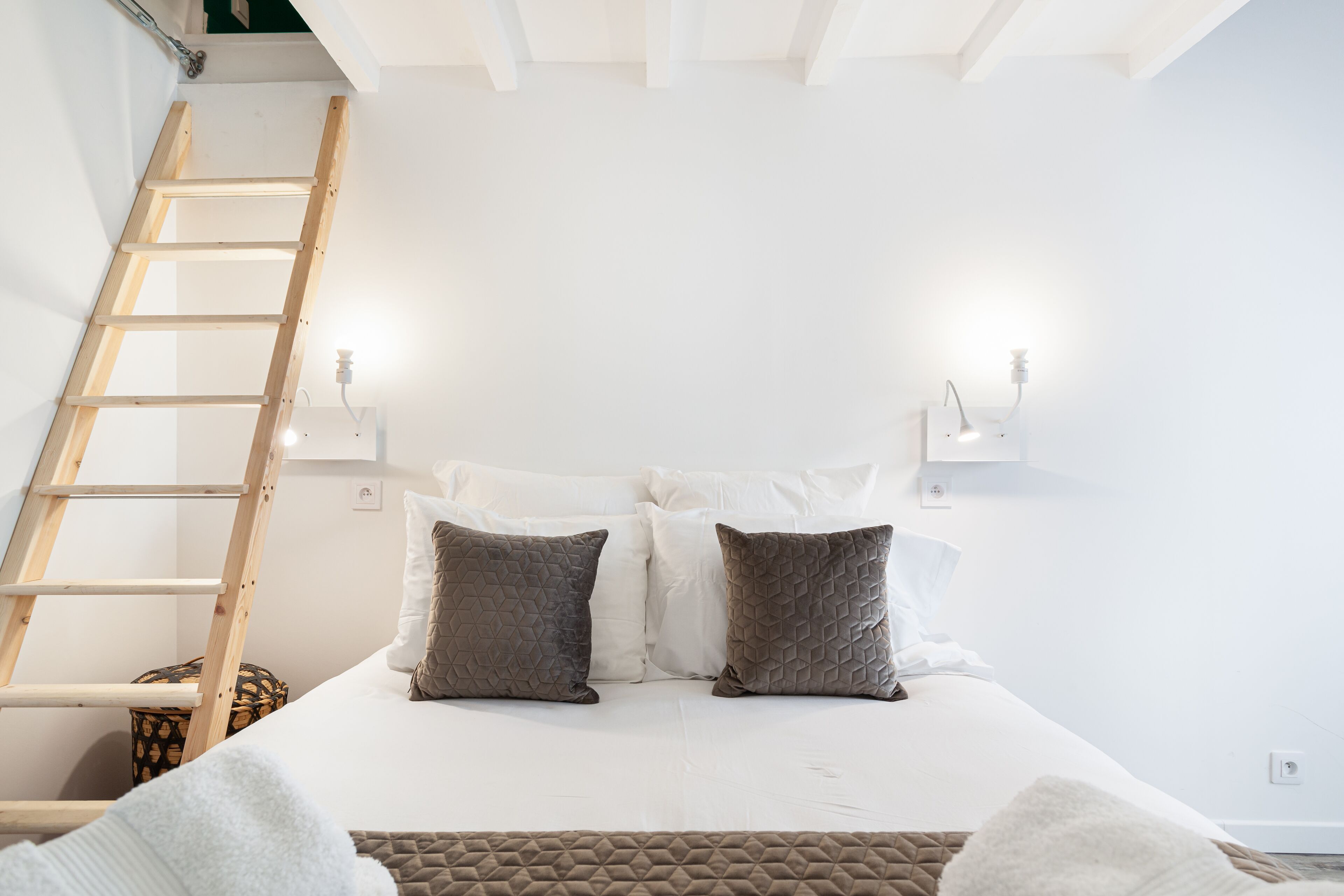 Un dormitorio con cama, almohadas, una escalera y luces fijadas a la pared.