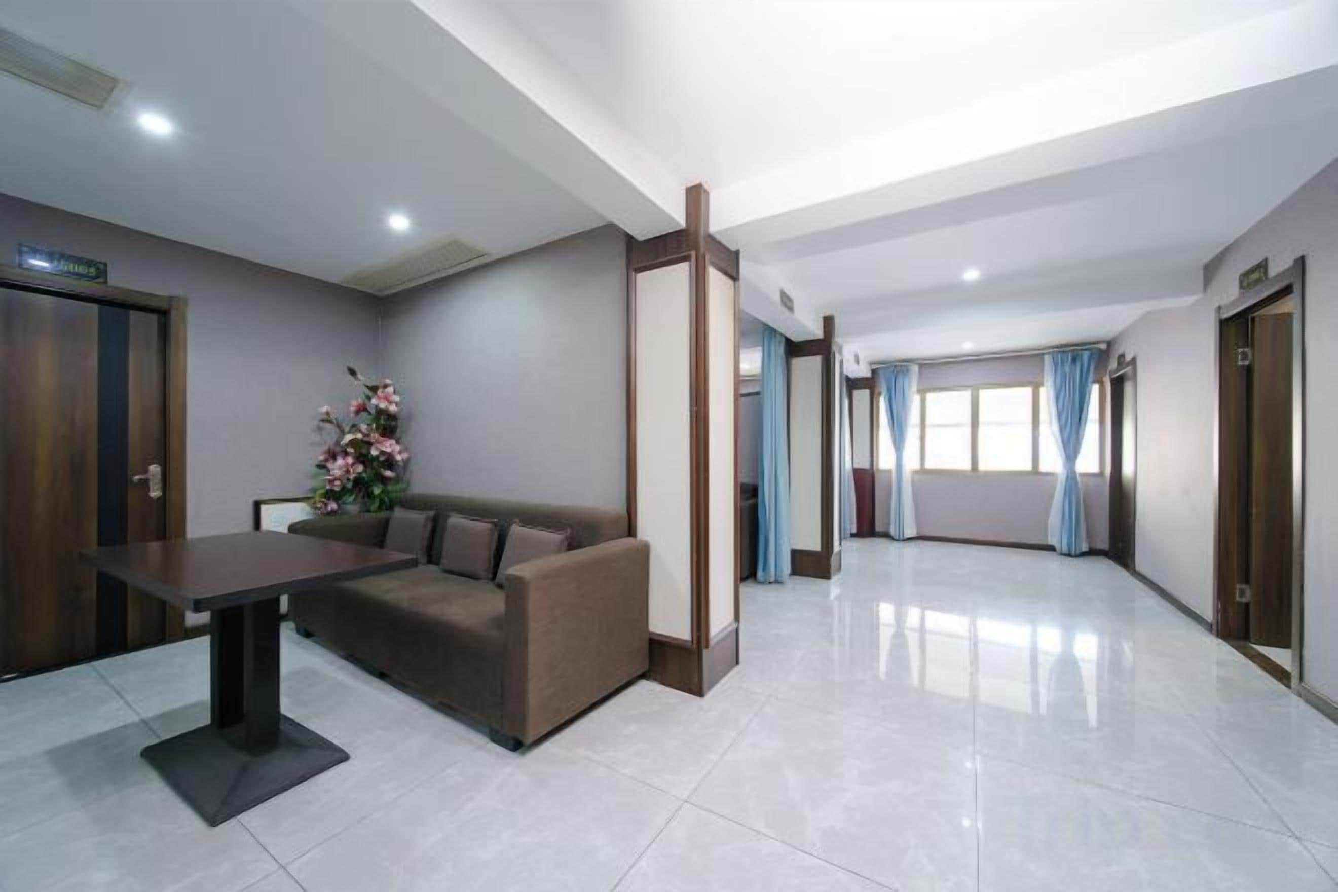 A hotel lobby with a reception desk, a sofa, a flower arrangement, and a corridor leading to elevators.